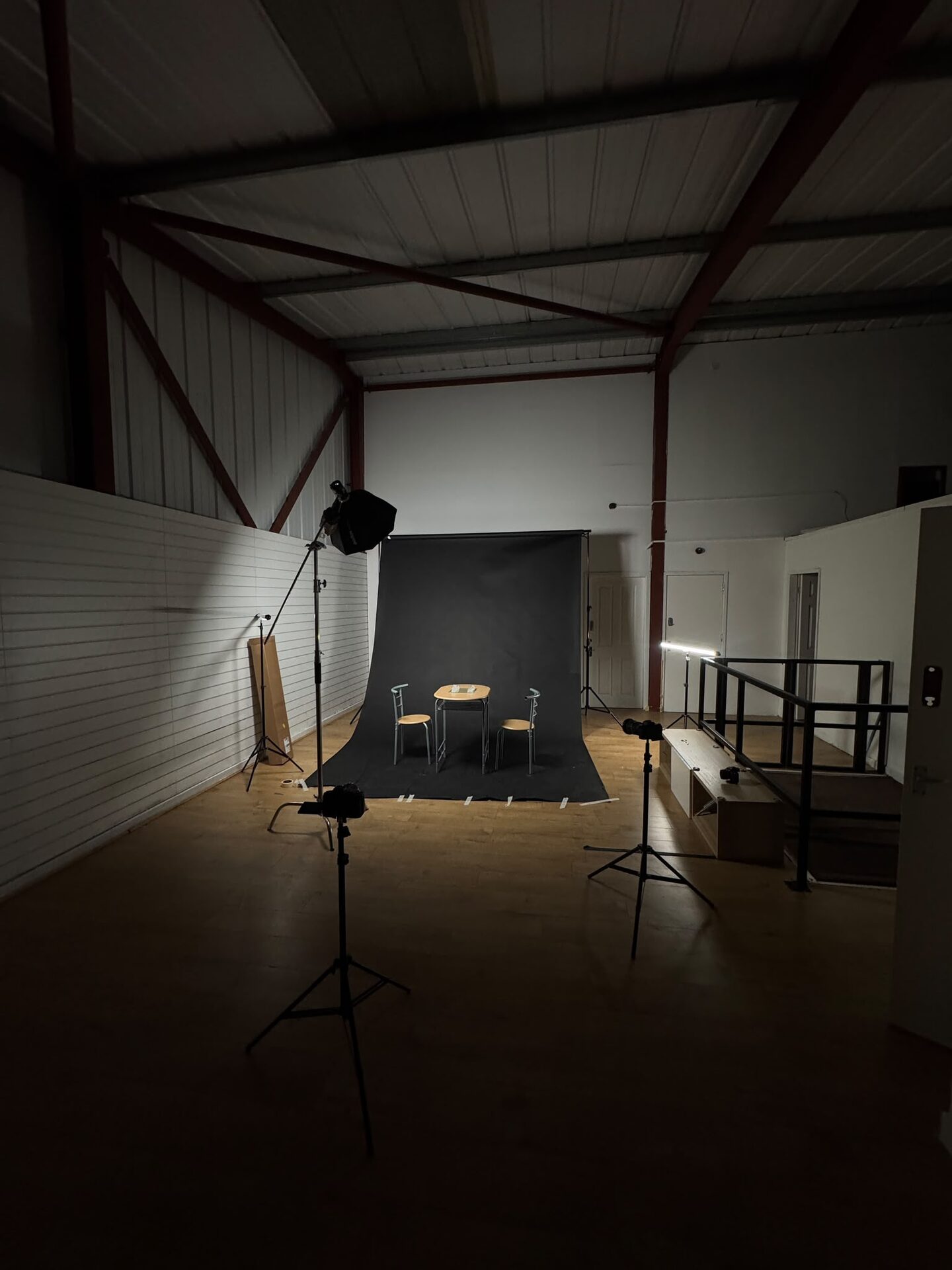 Empty studio with a black backdrop, a table and two chairs under a softbox, steel roof beams above